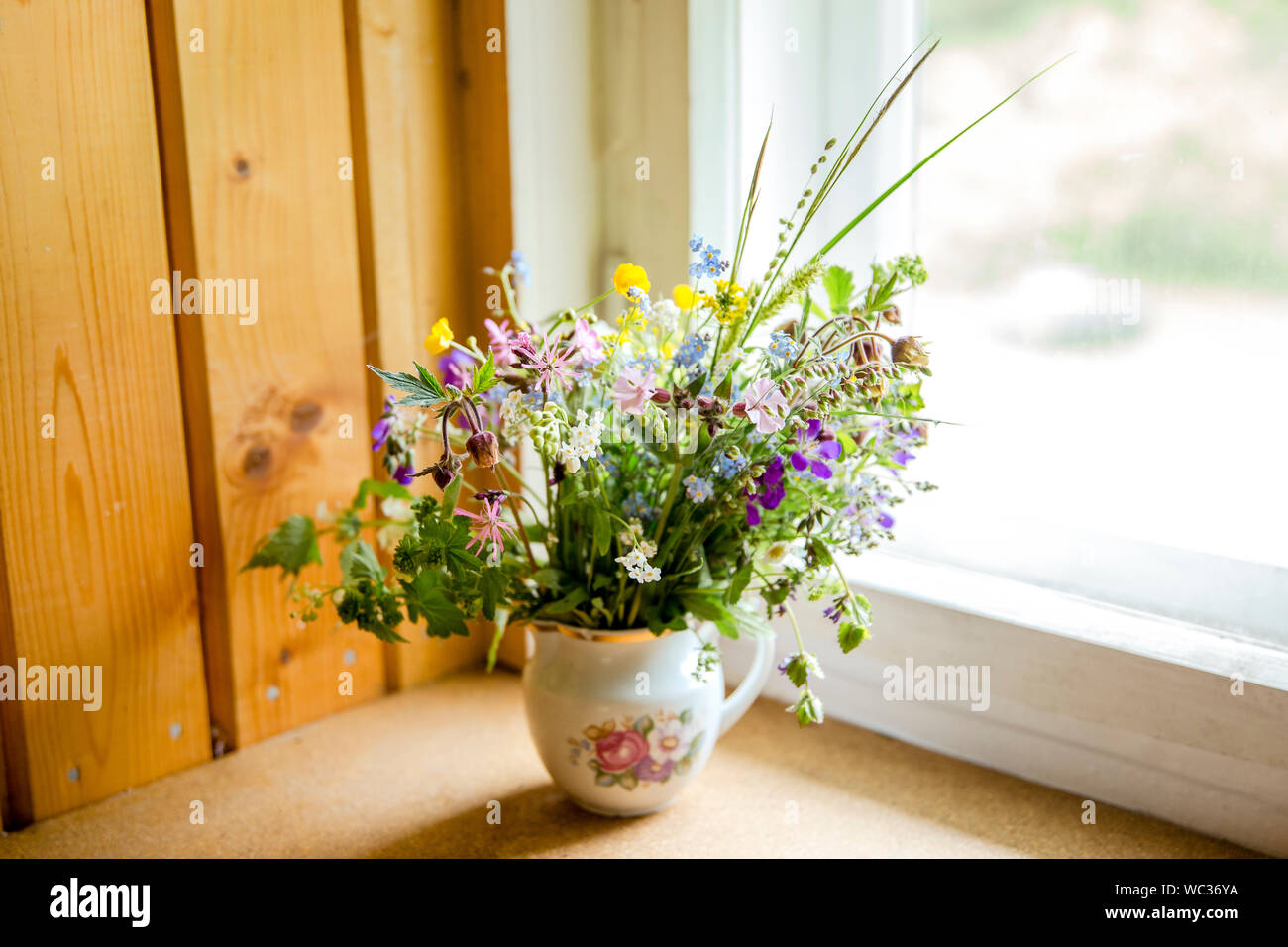 Vintage style flower bouquet made of wild flowers found in forest and meadow, standing in old cream jug on window sill, summers in grandma`s house con Stock Photo
